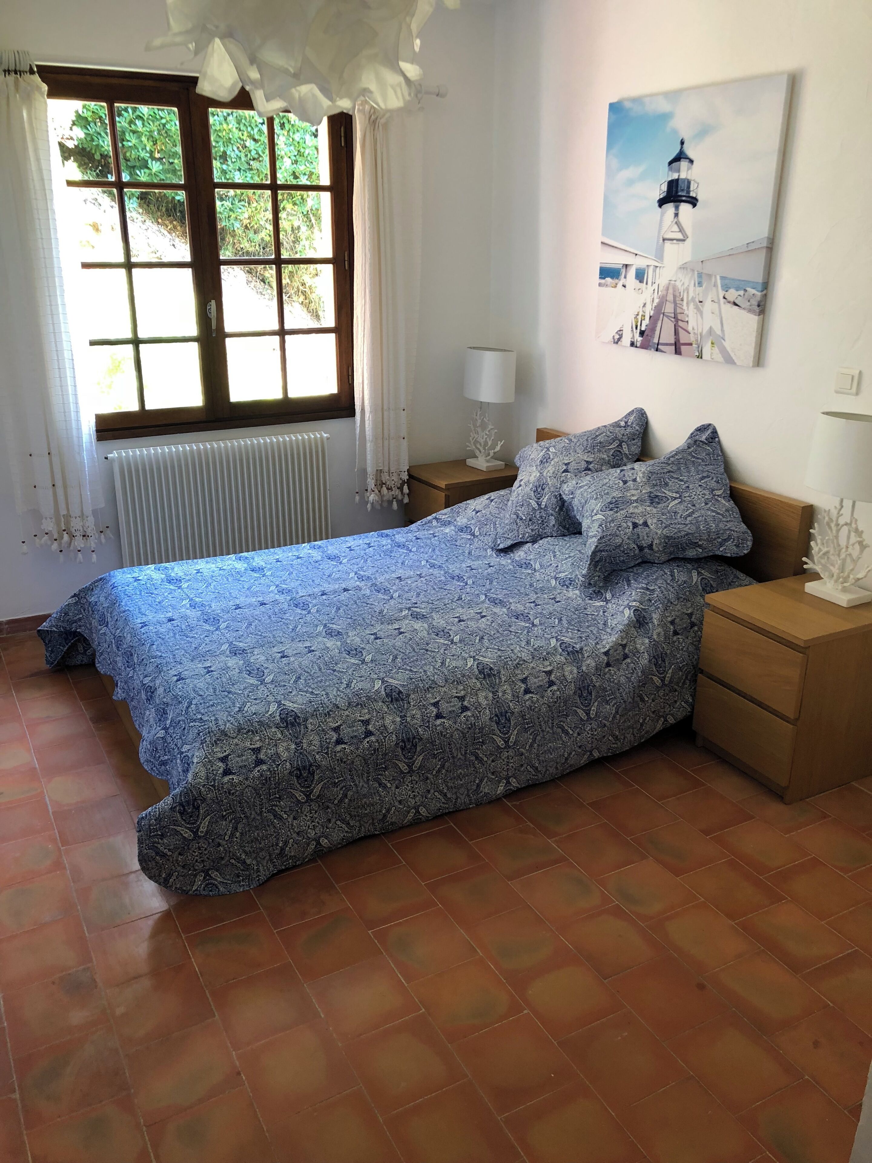 Une chambre à coucher comprenant un lit, une table de chevet, une lampe, une fenêtre avec des rideaux, un radiateur et un tableau accroché au mur.