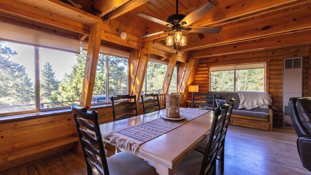 A wooden cabin interior with a dining table, chairs, a ceiling fan, and large windows offering a view of trees. - Enjoy a cozy cabin near the lake! Sleeps 8,  4 Beds. (Panguitch Lake)