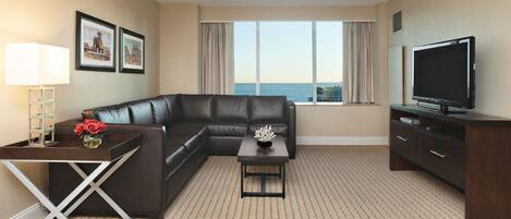 A hotel room with a sectional sofa, a small table with a vase of flowers, a TV, and a large window with a view of the sea.