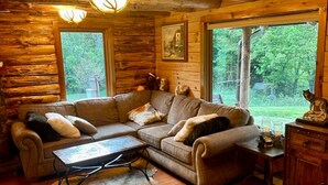 L'intérieur d'une cabane en rondins, avec un plancher en bois, un canapé en cuir, une table basse et une grande fenêtre donnant sur le paysage extérieur.