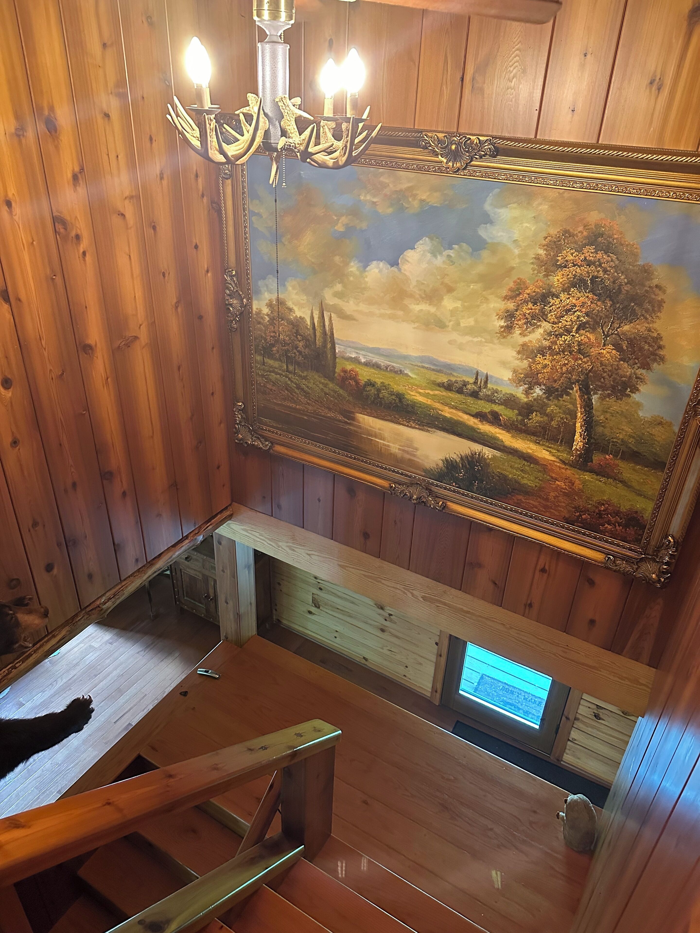 Un intérieur en bois orné d’un tableau représentant un paysage, d’un lustre et d’un escalier.
