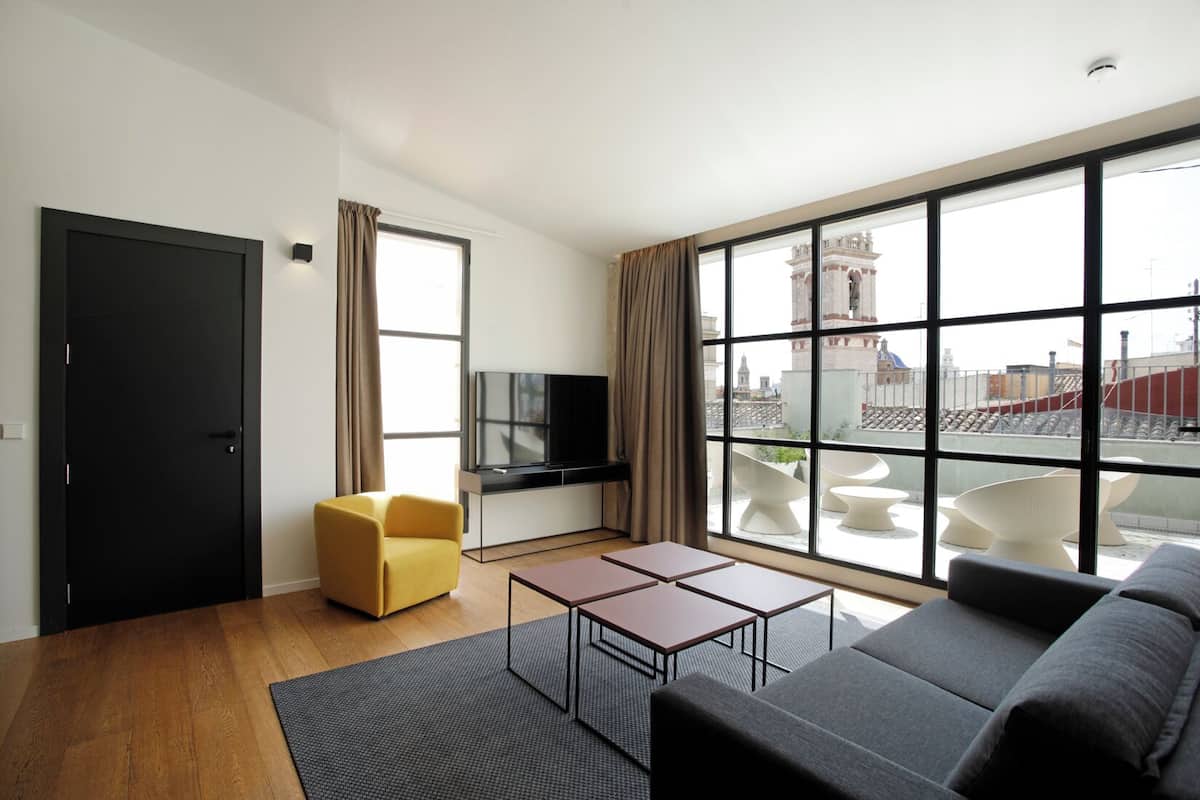 A modern living room with a sofa, a yellow armchair, a TV, and a large window with a city view.