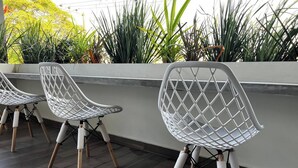 Una hilera de sillas blancas de estructura metálica y patas de madera, sobre un piso de baldosas oscuras, frente a una pared blanca con un macetero de plantas verdes al fondo.