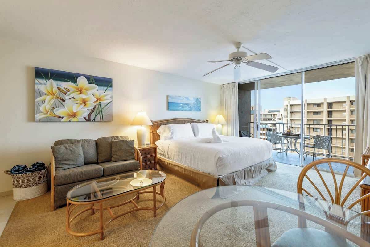 Een hotelkamer met een bed, een bank, een glazen tafel, een plafondventilator en een balkon met stoelen.