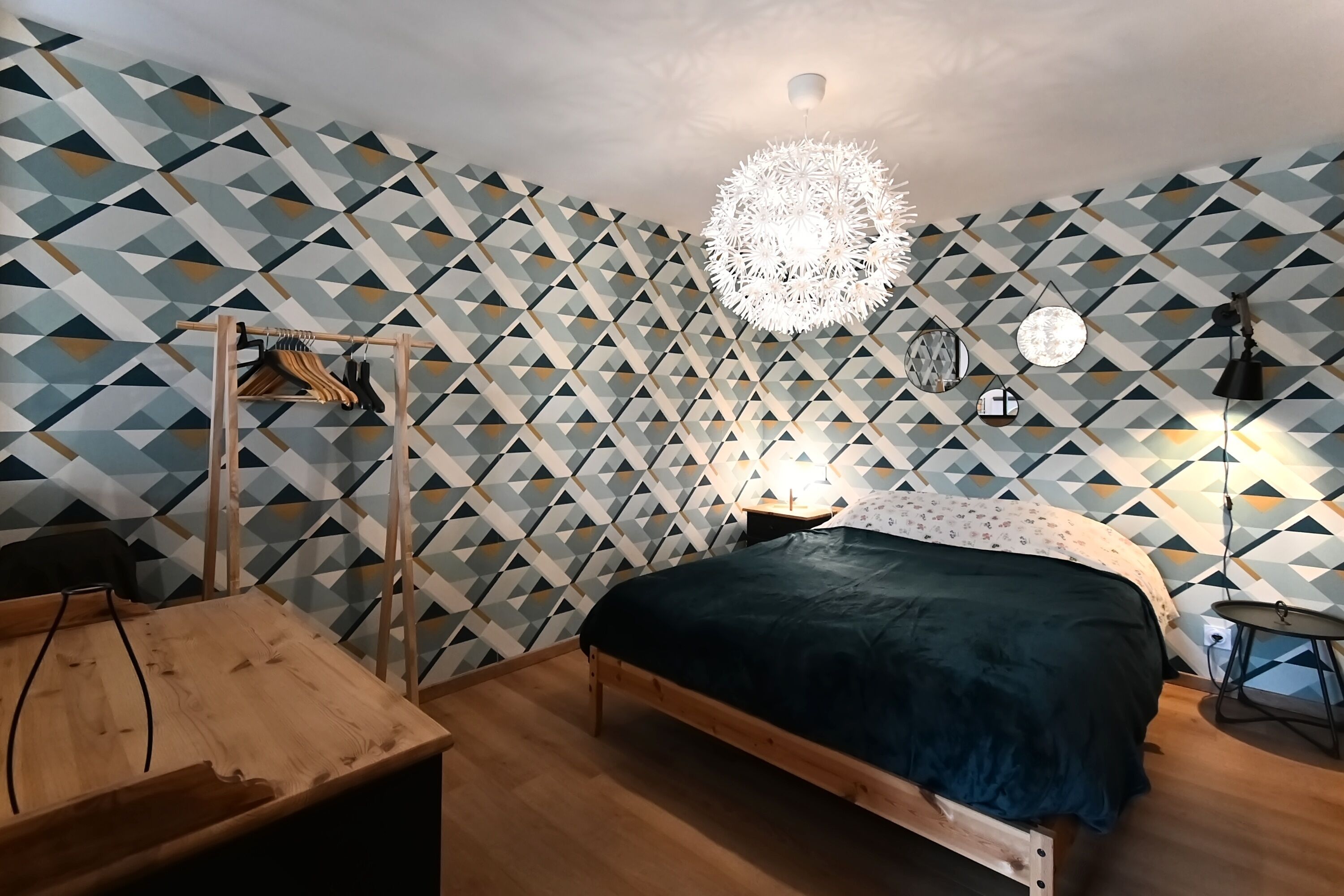 Une chambre avec un mur orné d’un motif géométrique, un lit recouvert d’une couvre-lit vert foncé, une table de chevet en bois, un luminaire suspendu et une petite table de chevet avec une lampe.