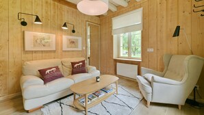 Une pièce chaleureuse avec des murs en bois, un canapé blanc, un fauteuil beige, une table basse en bois et deux lampes fixées au mur.