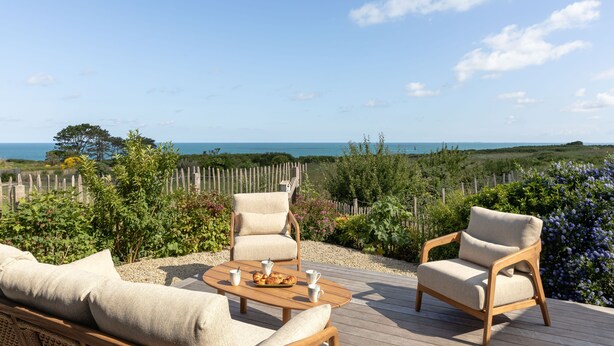 Eine Terrasse mit zwei beigen Sesseln, einem Holztisch mit einer Auswahl an Gebäck und einem Korbsessel.