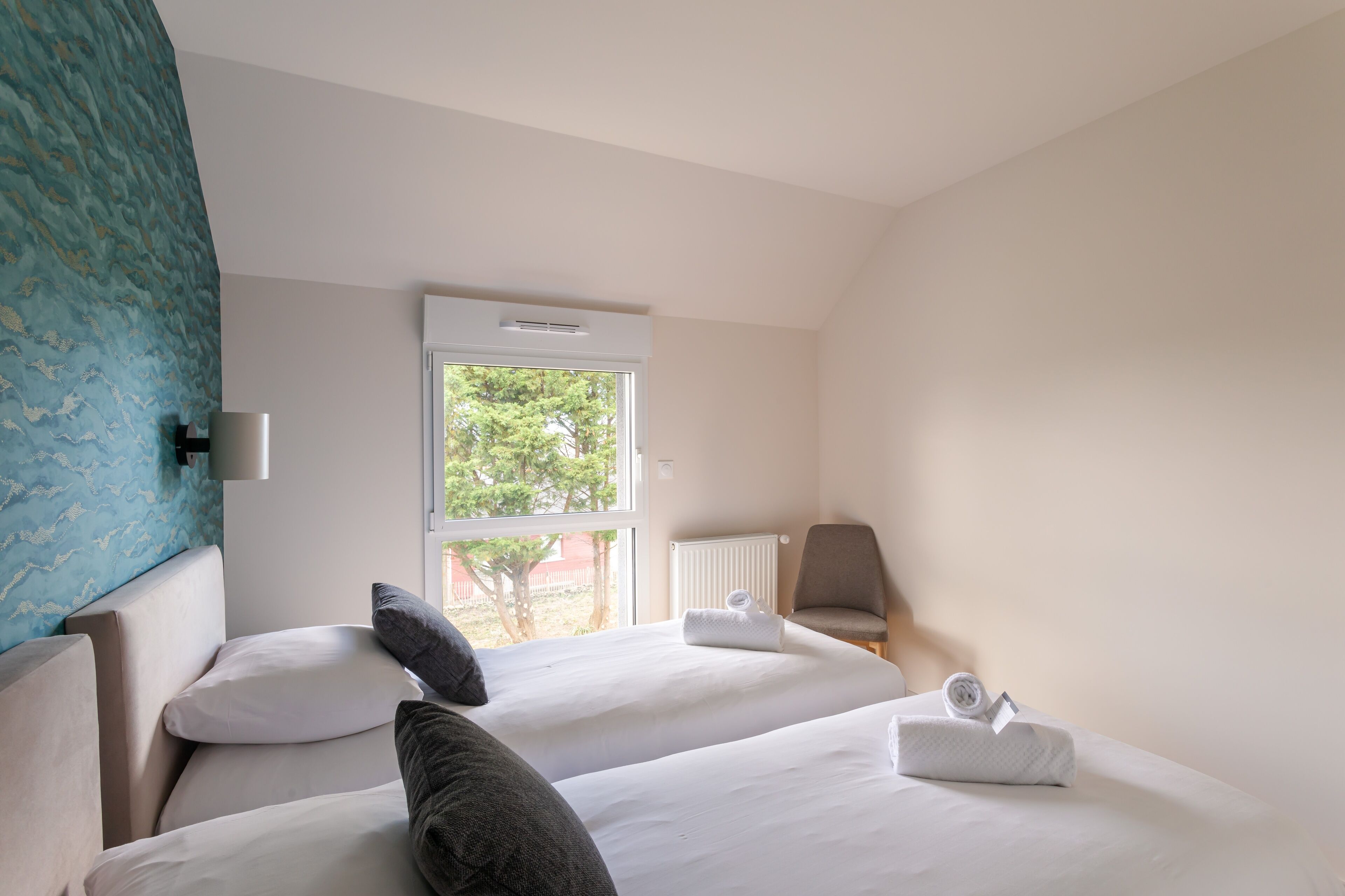 A hotel room with two beds, a window, a radiator, and a chair.