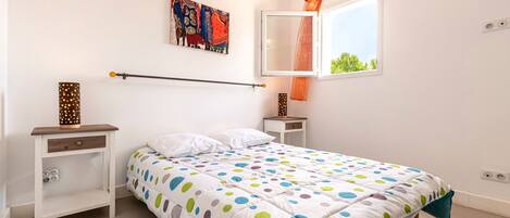 A bedroom with a bed, a nightstand, a wall-mounted shelf, a window with curtains, and a wall art piece.