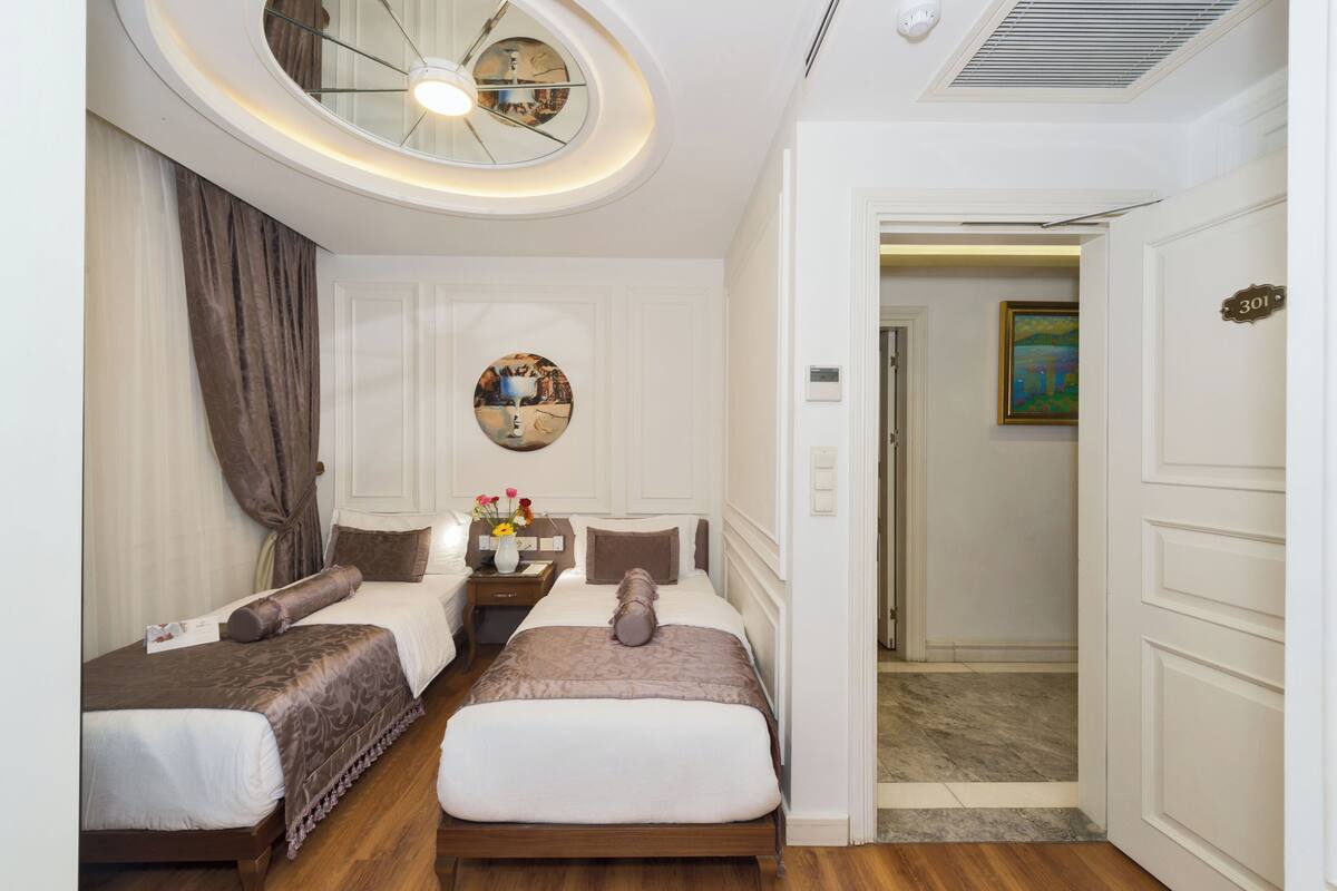 A hotel room with two beds, a round ceiling light, a painting on the wall, and a door with a number.