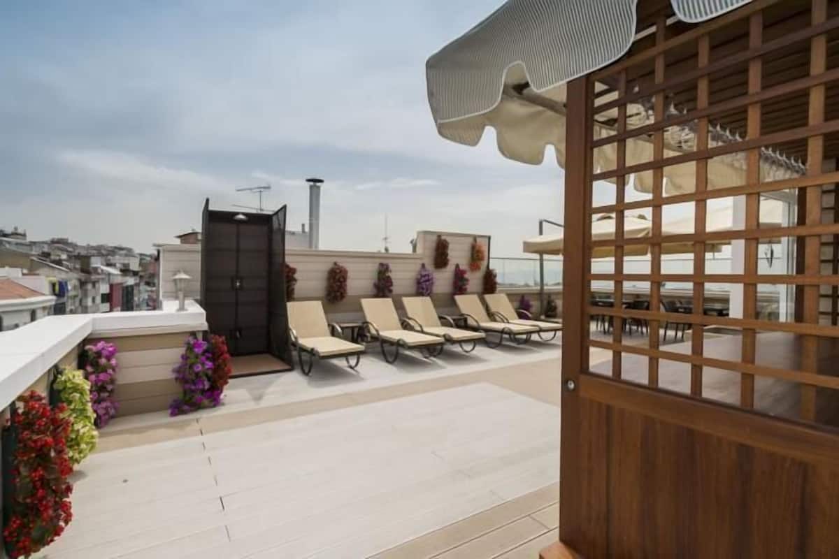 A rooftop terrace with sun loungers, a wooden gate, and a view of the cityscape.