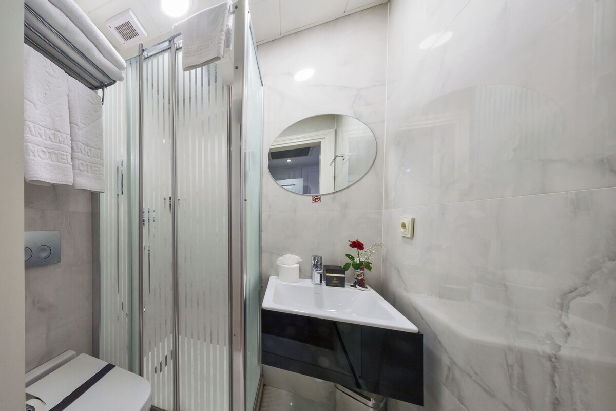 A modern bathroom with a glass shower enclosure, a toilet, a sink with a mirror, and a small trash bin.