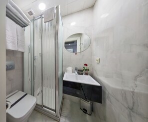 Egy modern fürőszoba, melyben üvegfalas zuhanyzó, WC, tükörrel ellátott mosdókagyló és egy kis szemeteskosár található.