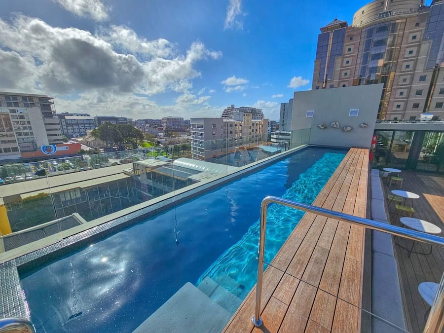 Une piscine à débordement située sur le toit, avec une terrasse en bois et des garde-corps en verre, offrant une vue panoramique sur la ville.