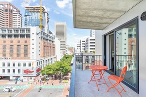 Un balcón con sillas naranjas, con vistas a una calle de la ciudad y edificios, incluyendo un restaurante 'Tiger' y un edificio 'Luno'.