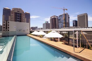 Une piscine sur le toit, dotée d’une terrasse en bois et de parasols, entourée de bâtiments modernes et surplombée par une grue de construction.