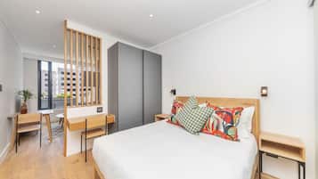 A modern bedroom with a large bed, a desk, and a chair. There is a window with a view of buildings outside.