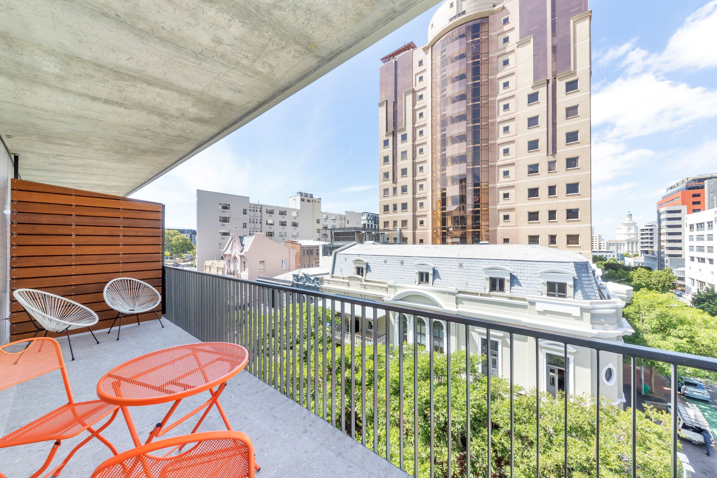 A balcony with orange metal chairs and a wooden slatted wall, overlooking a cityscape with various buildings and greenery.
