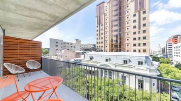 A balcony with orange metal chairs and a wooden slatted wall, overlooking a cityscape with various buildings and greenery.