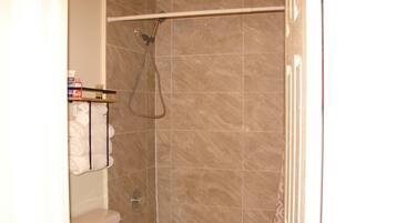 A bathroom with beige tiles, a shower with a handheld showerhead, a toilet, and a towel rack with folded towels.