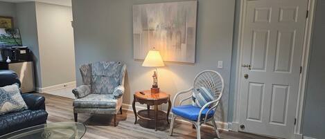 A living room with a blue armchair, a wooden side table with a lamp, a glass coffee table, and a black leather sofa.
