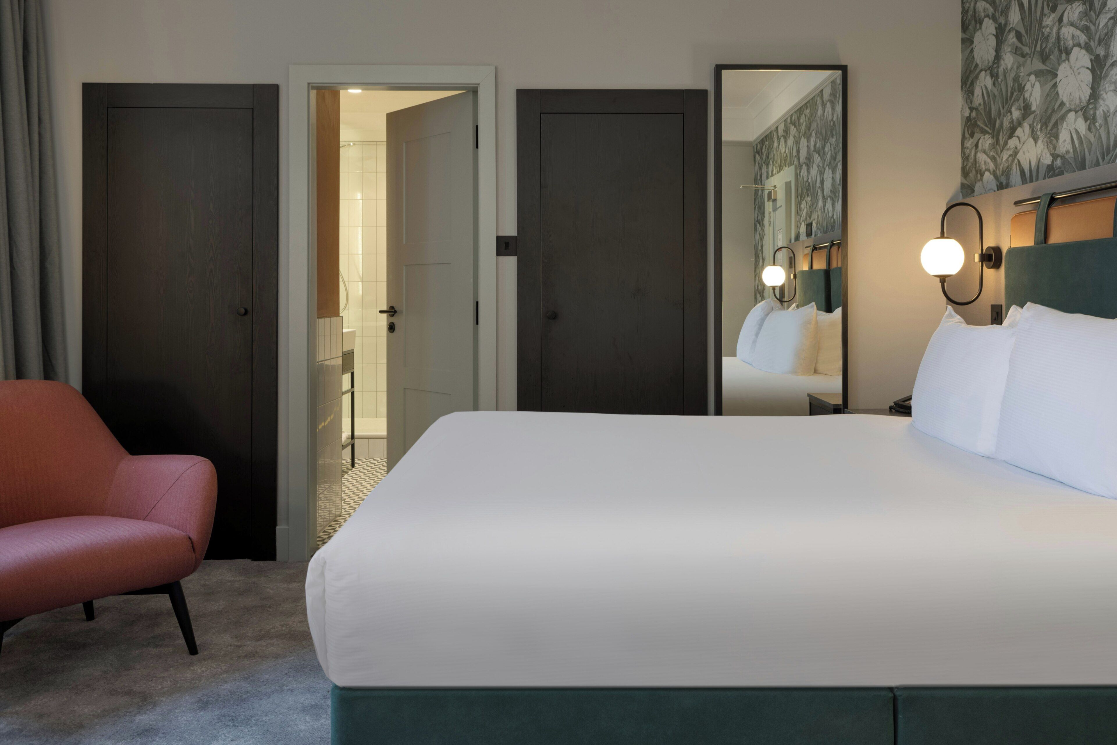 A hotel room with a bed, a chair, a mirror, and a door leading to a bathroom.