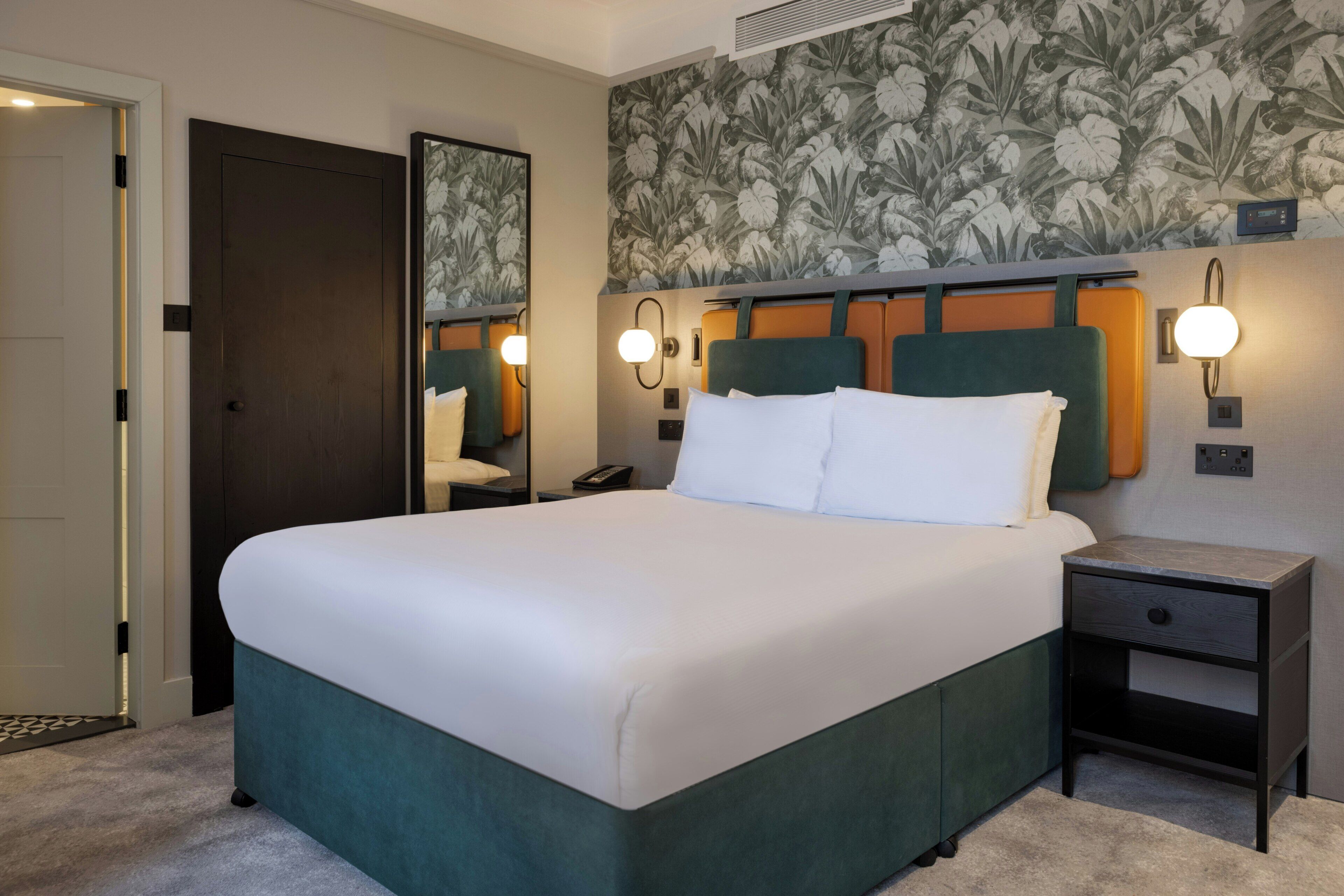 A hotel room with a large bed, a nightstand, a mirror, and a floral wallpaper.