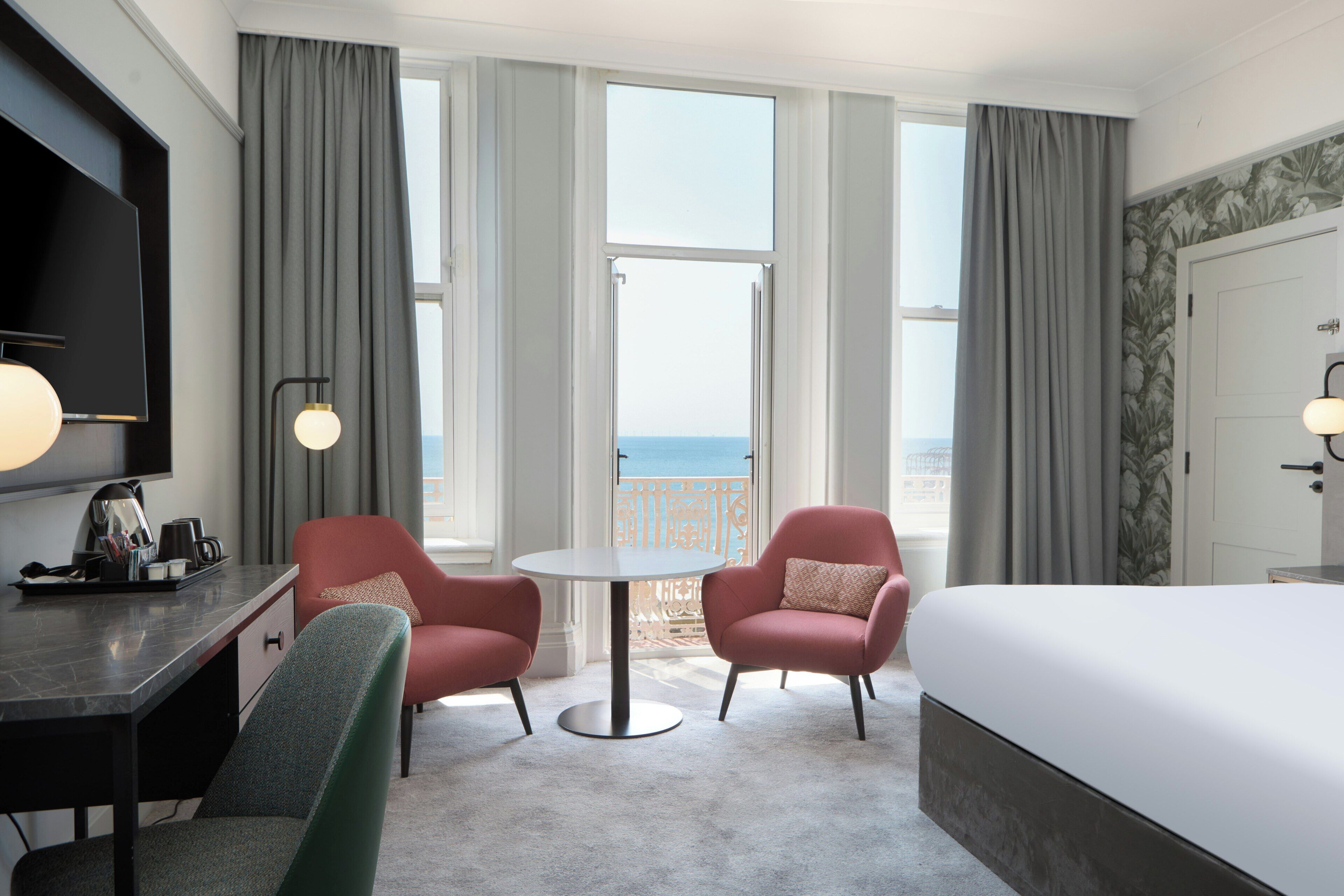 Habitación de hotel con cama, dos sillones, escritorio con televisor de pantalla plana, mesa redonda y ventanas amplias con vista al mar.