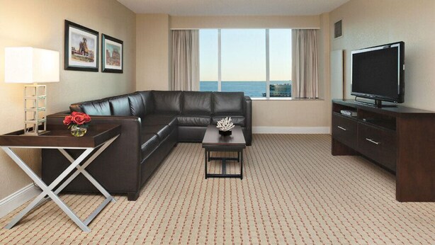Habitación de hotel con sofá modular, una mesita con un florero, un televisor y un ventanal con vista al mar.