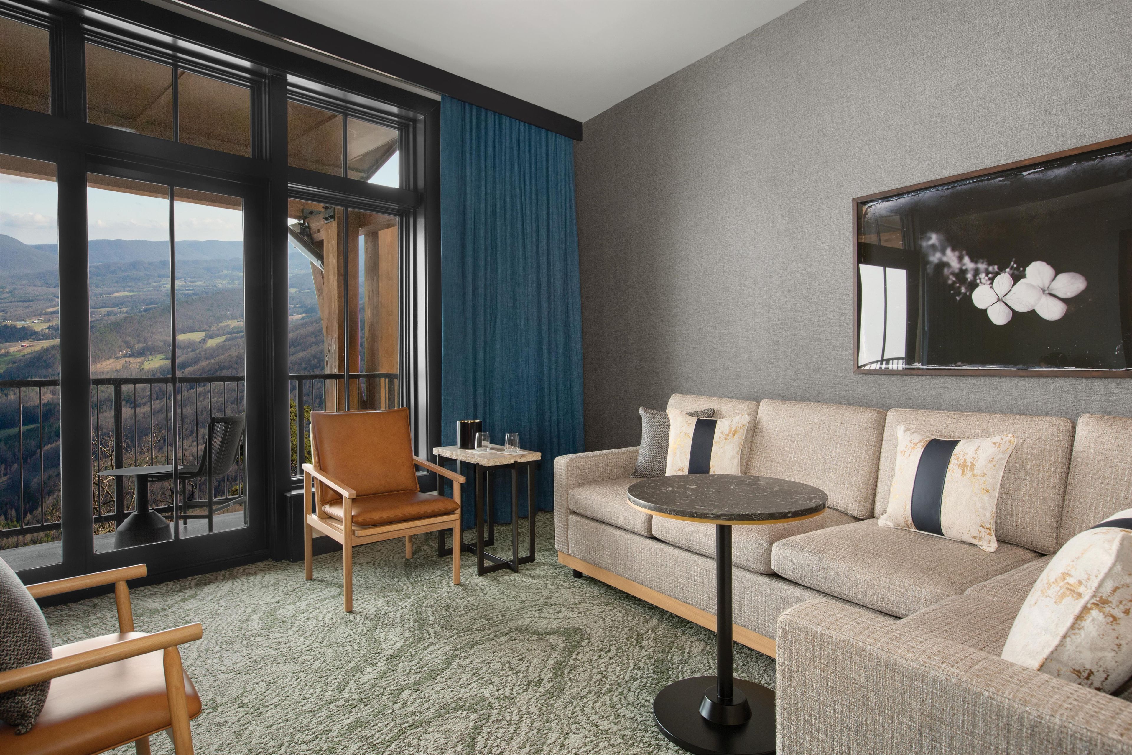 A modern hotel room with a large window offering a scenic view, a cozy seating area with a sofa and armchairs, and a small round table.