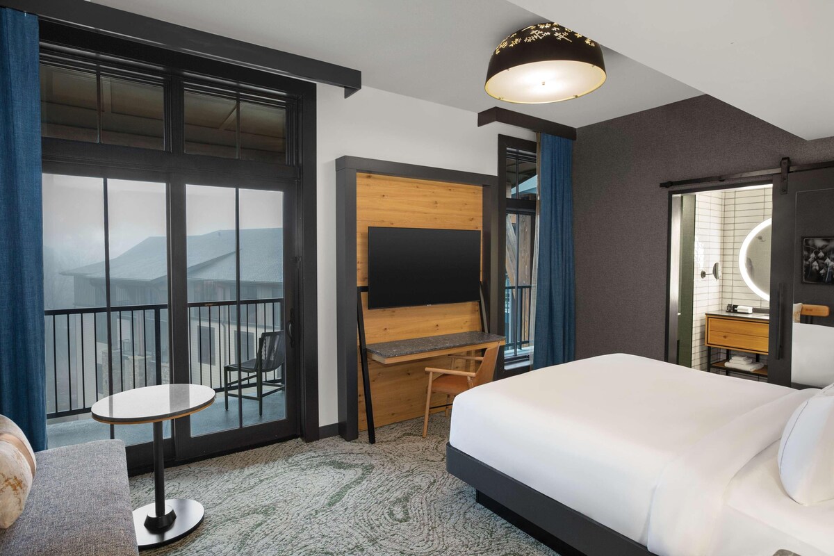 Une chambre d’hôtel comprenant un lit, un bureau, une chaise, une télévision et un balcon avec vue sur les montagnes.