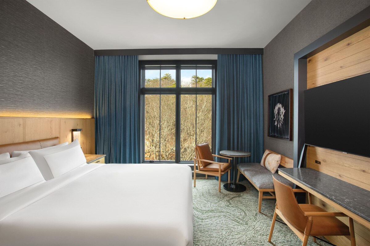 Une chambre d’hôtel avec un grand lit, un bureau, une chaise, une petite table et une fenêtre avec des rideaux.