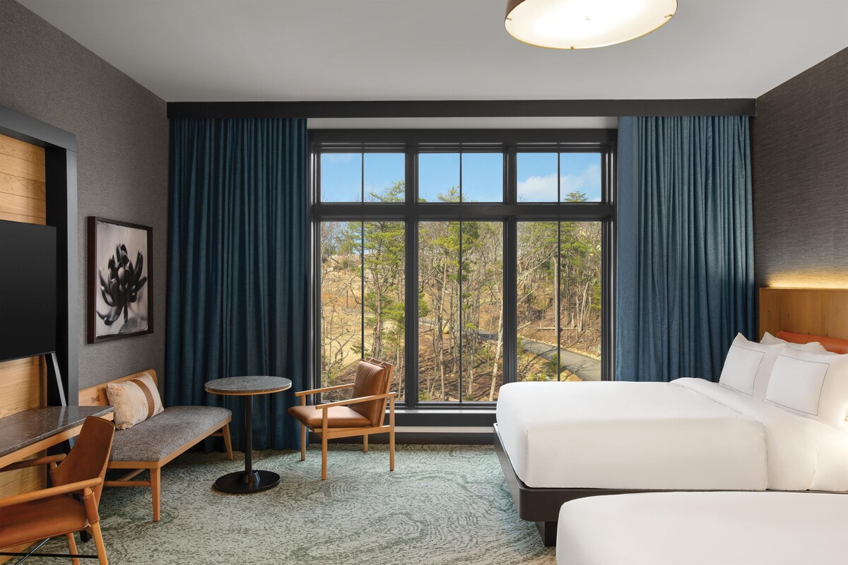 Une chambre d’hôtel avec une grande fenêtre, un lit, un bureau avec une chaise et une petite table ronde.