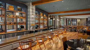 A bar area with wooden shelves stocked with bottles, a long counter, and several wooden chairs.