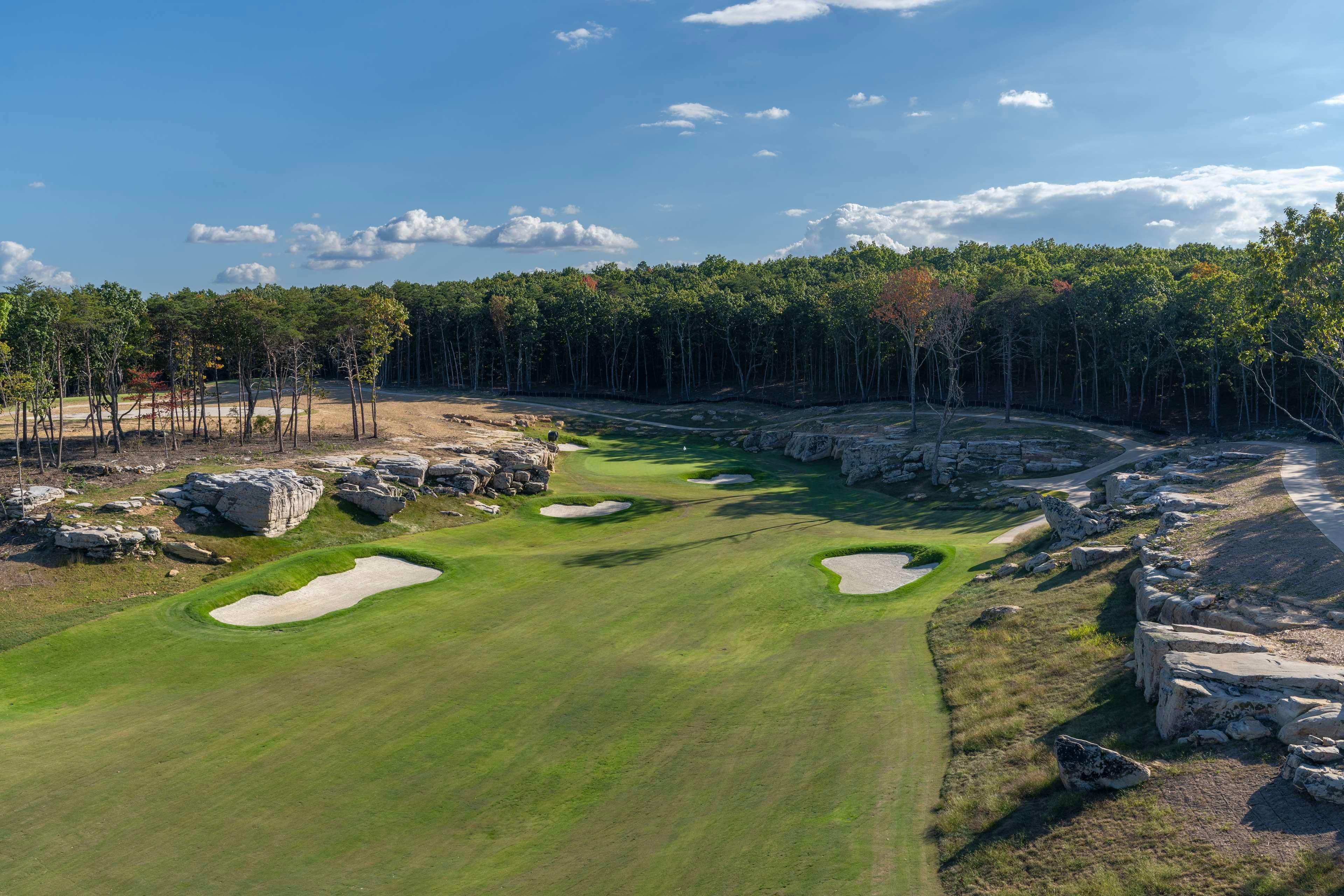 Egy golfpálya, melynek része a homokakadályokkal tarkított terület, tiszta az égbolt, és a háttérben erdő látható.
