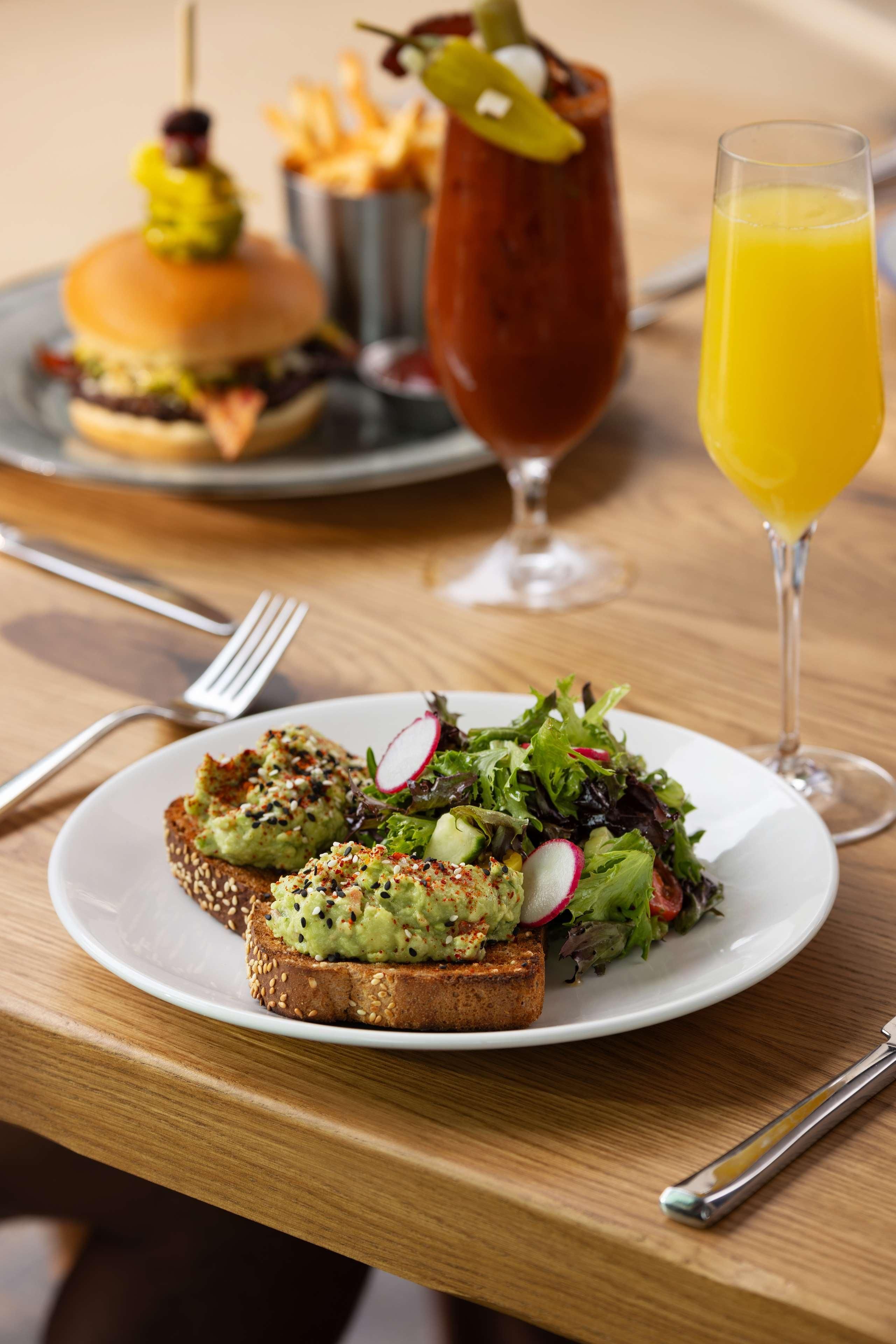 A plate with avocado toast and a salad, a burger, and a glass of orange juice on a wooden table.