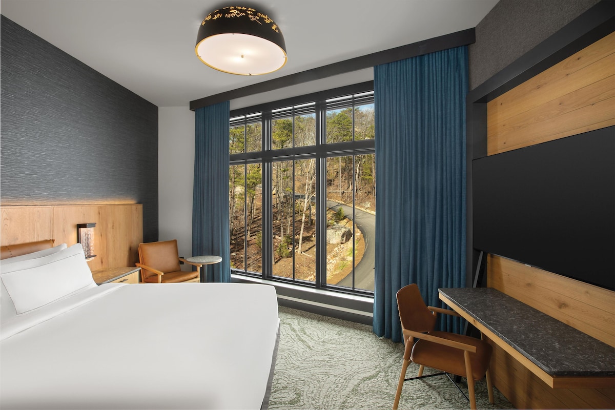 A hotel room with a bed, desk, chair, and large windows offering a view of a forested area.