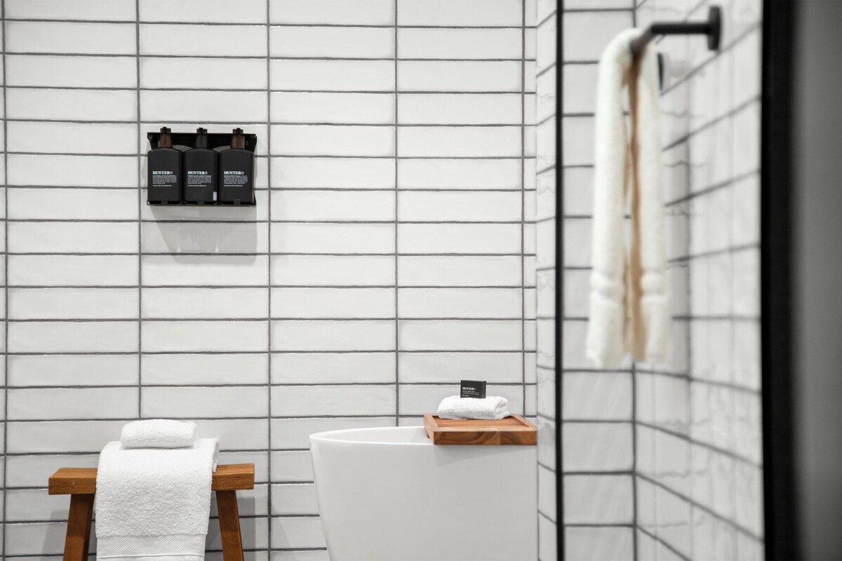 Une salle de bain avec un mur recouvert de carreaux blancs, un porte-serviettes en bois, un lavabo blanc et un distributeur de savon fixé au mur.