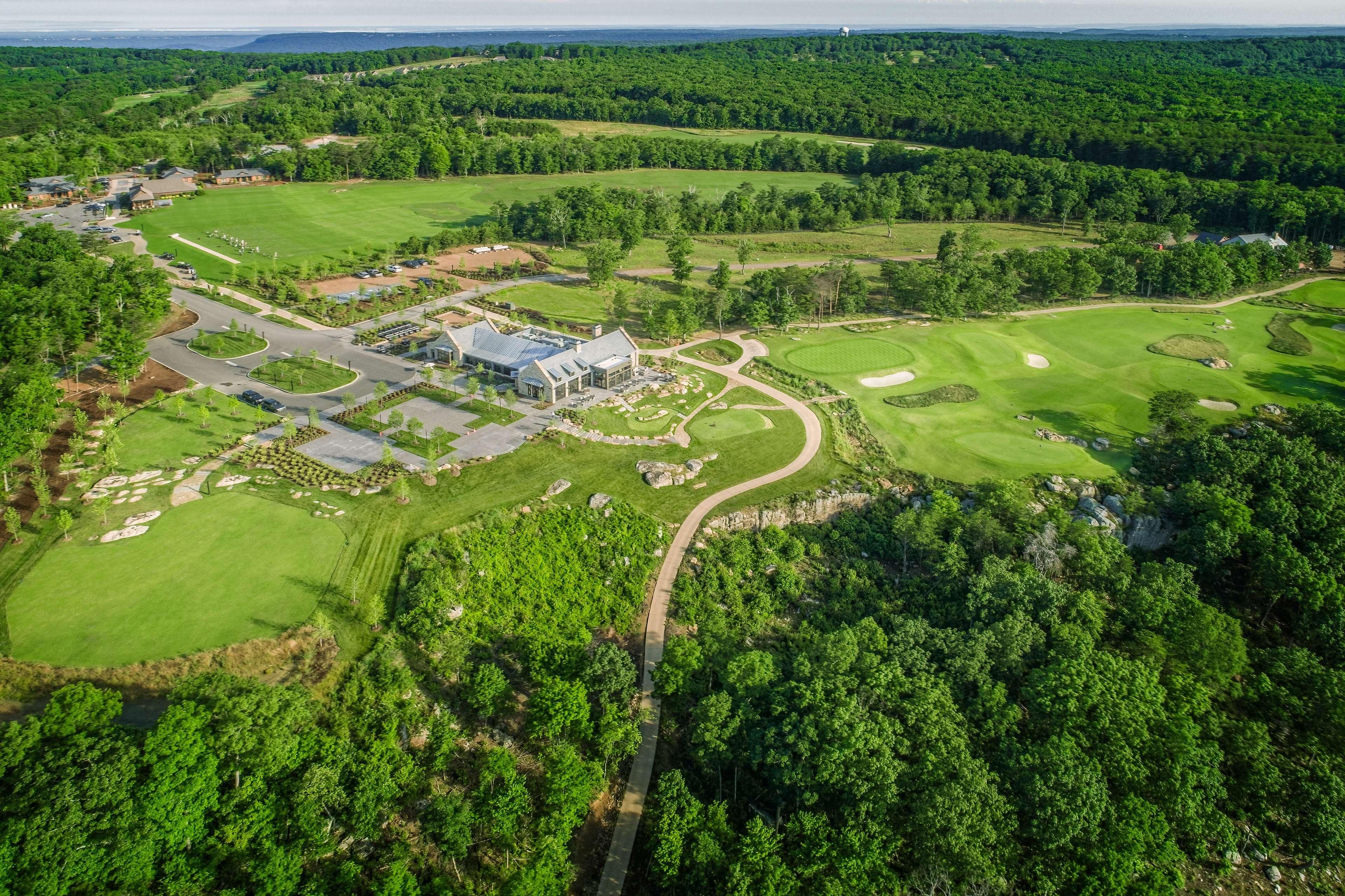 Vue aérienne d’un terrain de golf avec un club-house, plusieurs greens et des zones boisées environnantes.