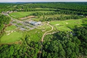 Vista aérea de un campo de golf con un club de campo, múltiples áreas verdes y zonas boscosas circundantes.
