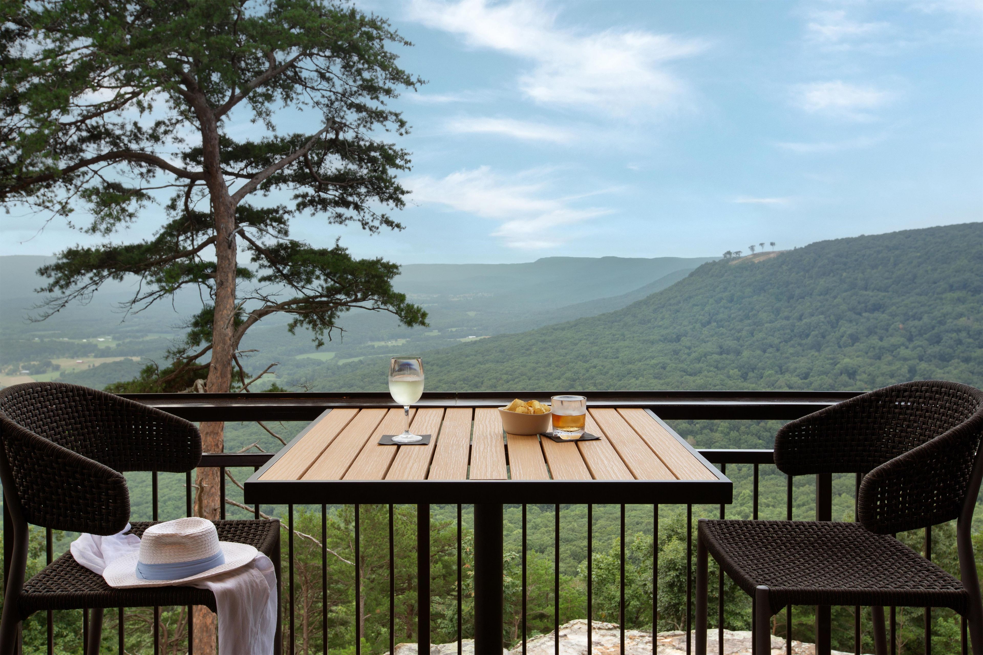 Una mesa de madera con dos sillas de mimbre, un sombrero y una copa de vino en un balcón con vistas a un paisaje montañoso.