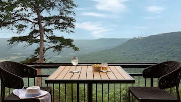 A wooden table with two wicker chairs, a hat, and a glass of wine on a balcony overlooking a mountainous landscape.