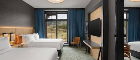Hotelkamer met twee bedden, een bureau, een stoel, een televisie en een groot raam met gordijnen.