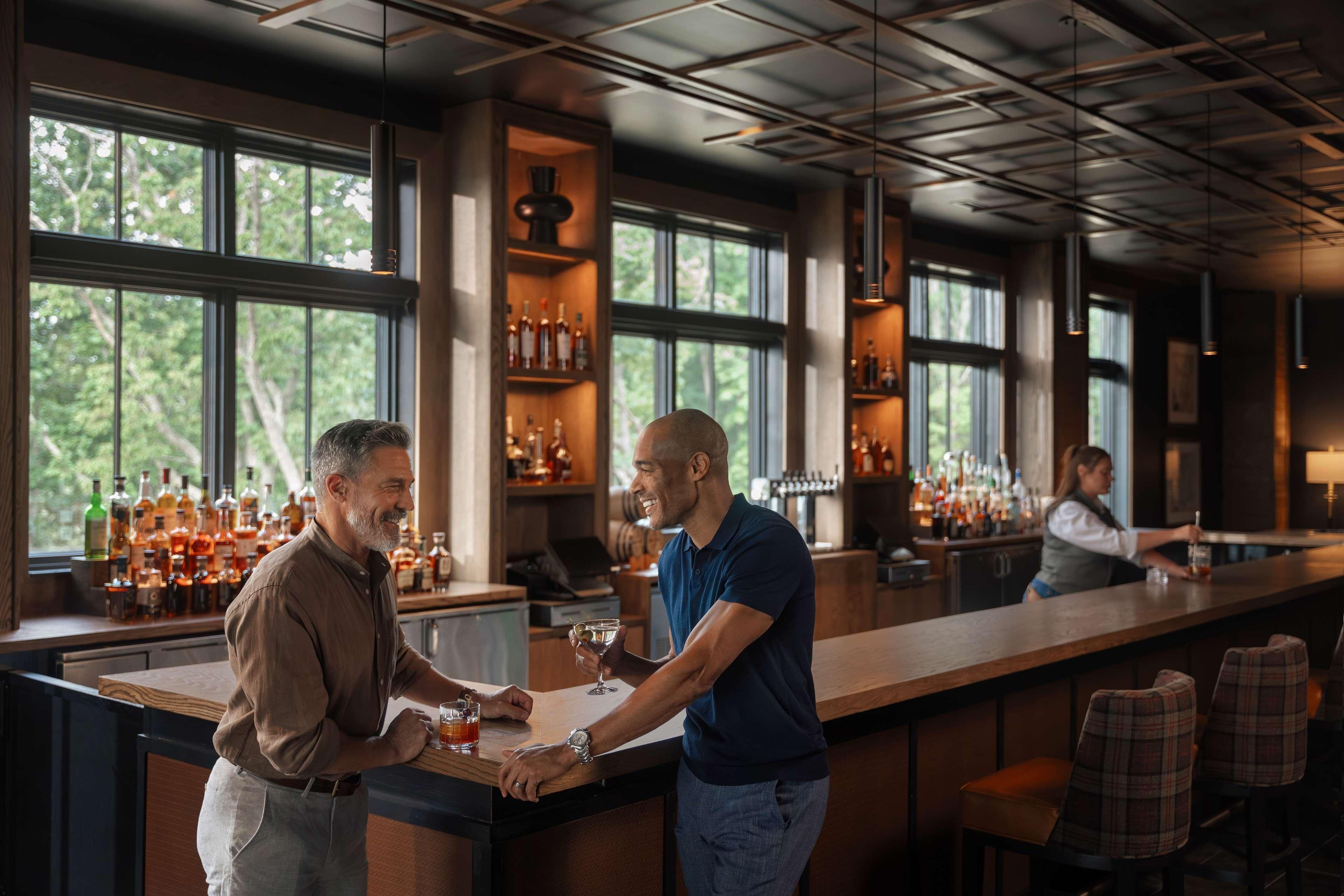 A bar area with a long counter, multiple shelves of bottles, and a view of trees through large windows.
