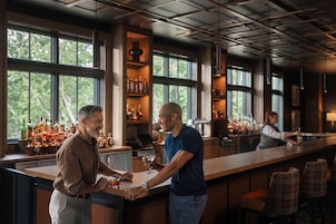 A bar area with a long counter, multiple shelves of bottles, and a view of trees through large windows.