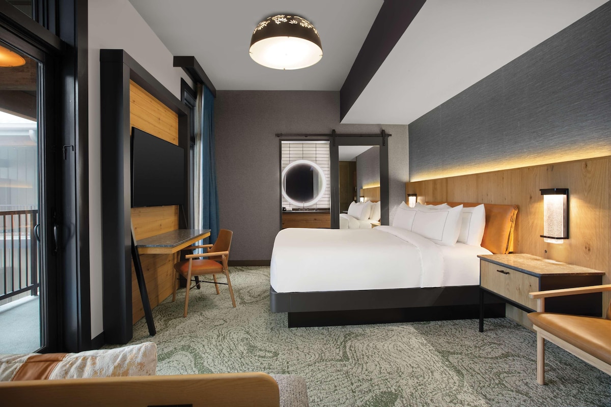 A modern hotel room with a large bed, a desk, and a chair. There is a window with a view of the outside.