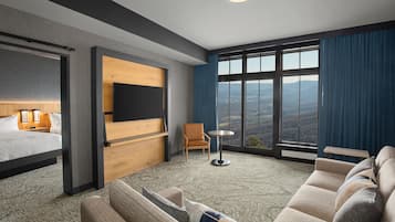 A hotel room with a bed, a seating area with a sofa and armchairs, a small table, a TV mounted on a wooden panel, and a large window offering a view of the mountains.