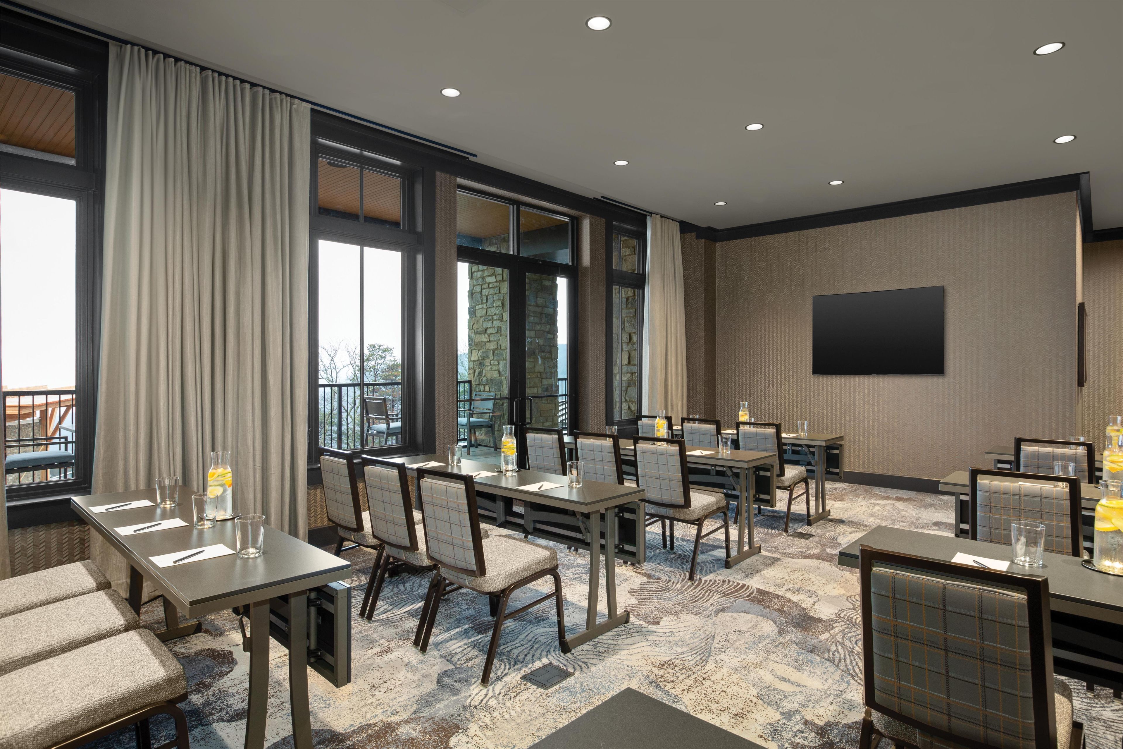 A hotel conference room with multiple tables and chairs, large windows, and a flat-screen TV on the wall.