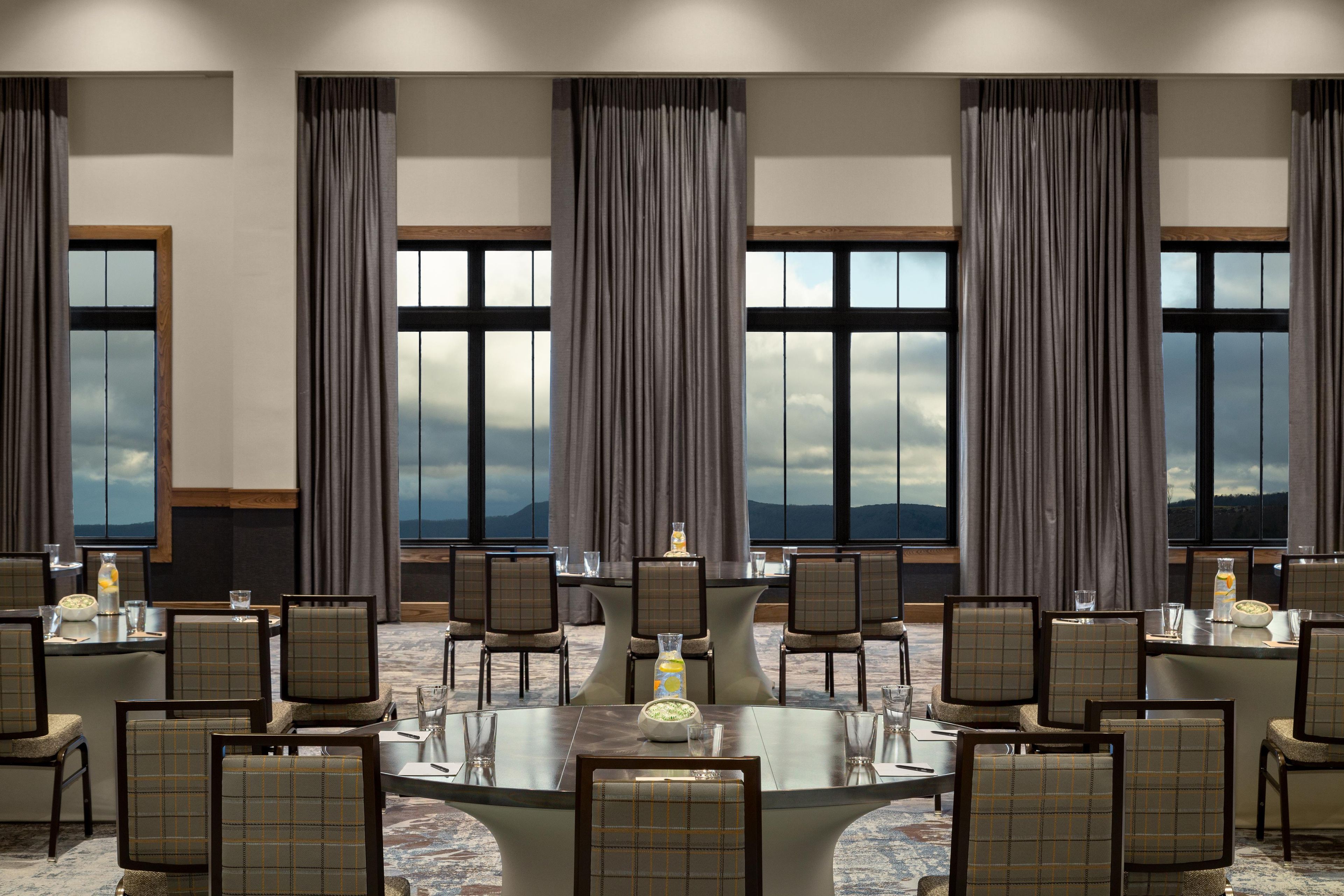 A conference room with a round table, chairs, and large windows with curtains.