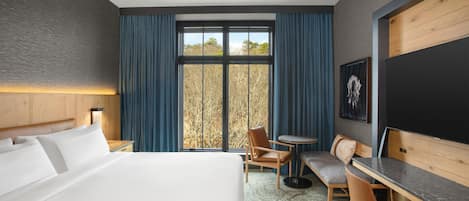 Ein Hotelzimmer mit einem großen Bett, einem Schreibtisch, einem Sessel, einem kleinen Tisch und einem Fenster mit Vorhängen.